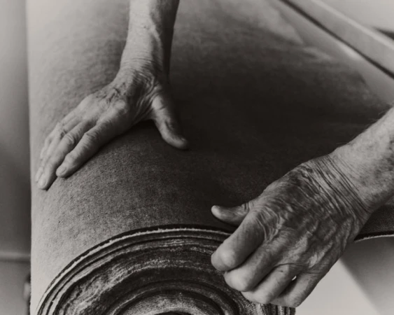 Close-up monochrome image of elderly hands unrolling a thick roll of textured fabric material.