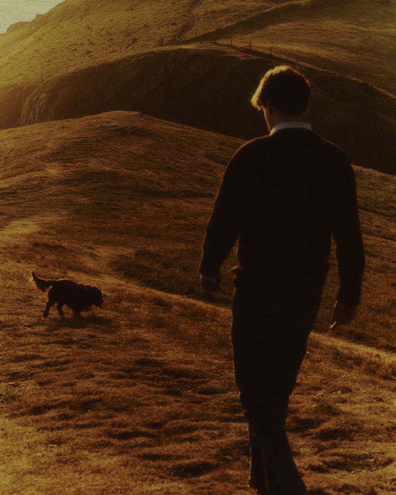 Man walking with a dog on a grassy hillside under warm golden hour lighting photographed by Max Miechowski