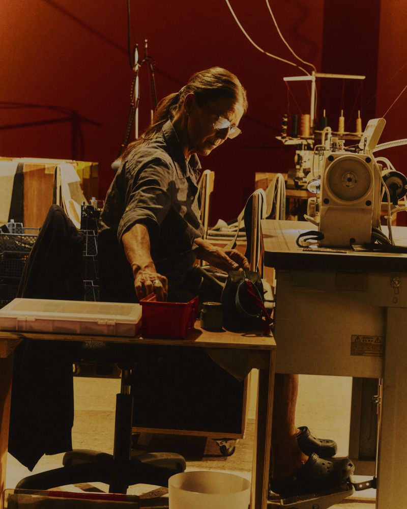 An older woman seated at a workbench in a workshop working with sewing equipment.