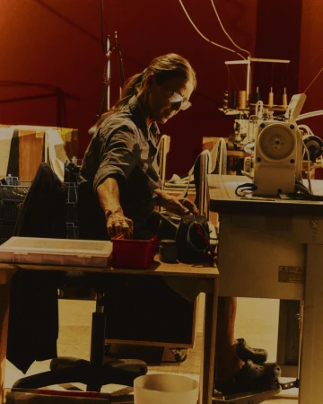 An older woman seated at a workbench in a workshop working with sewing equipment.