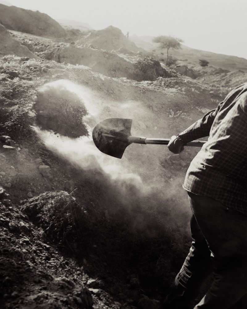 Monochrome editorial image of a person digging earth with a shovel, kicking up dust in a sparse landscape for Atmos Magazine.