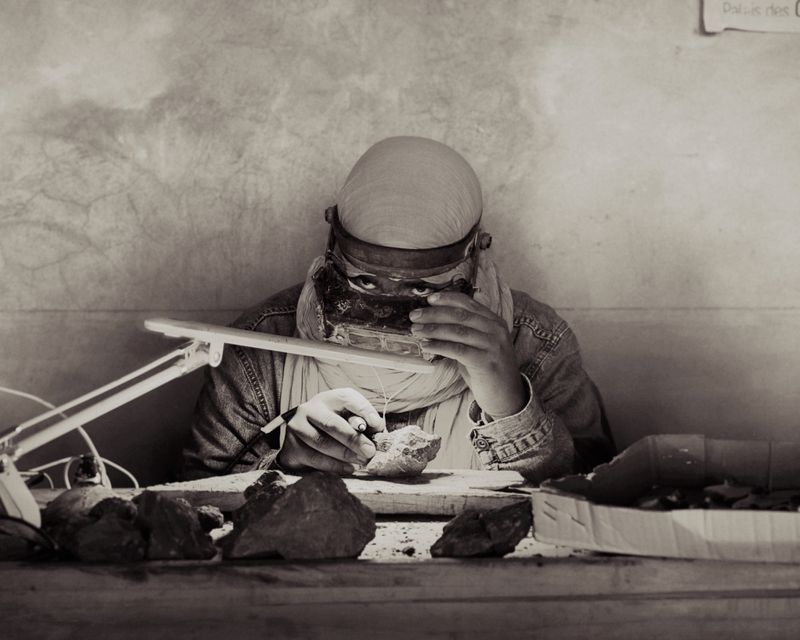 Person examining a rock specimen with a magnifying glass under a desk lamp in a monochromatic documentary photograph