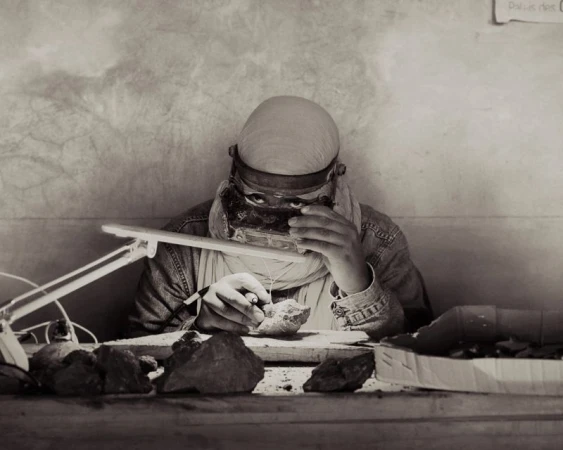 Person examining a rock specimen with a magnifying glass under a desk lamp in a monochromatic documentary photograph