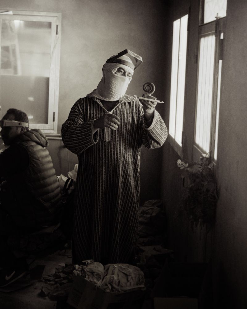 Man wearing a striped tunic and face covering holding an object inside a room with bright windows, photographed by Max Miechowski.