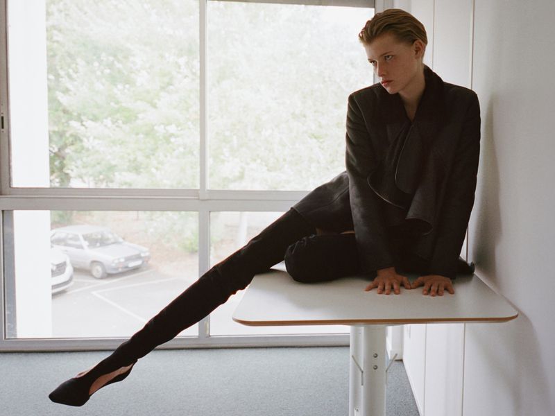 A model seated on a high white table near a window, wearing a dark jacket and trousers, with one leg extended.