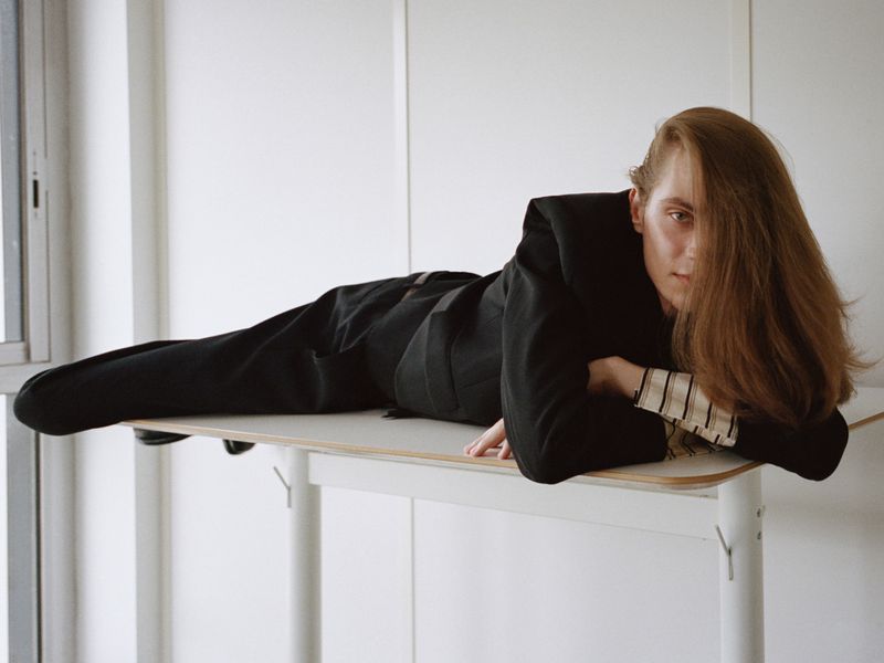 Model lying horizontally on a white standing desk wearing a black blazer and striped shirt for AnOther Magazine editorial.