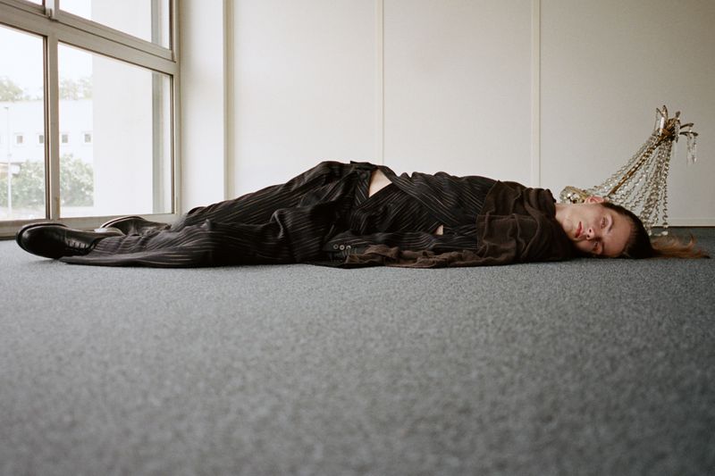 Model lying horizontally on a grey carpeted floor near a large window wearing a dark pinstripe suit