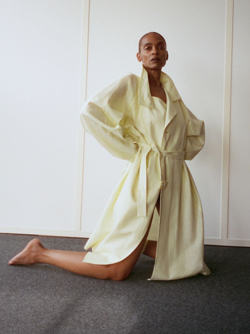 Model kneeling on dark carpet wearing a pale yellow belted coat against a plain white wall