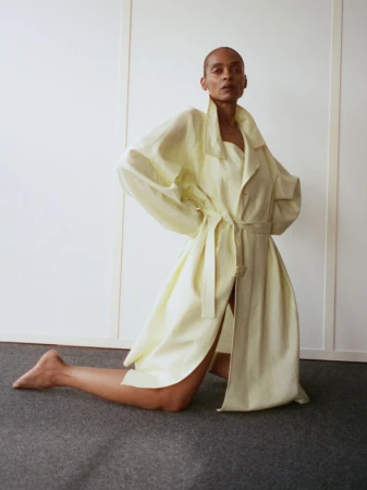 Model kneeling on dark carpet wearing a pale yellow belted coat against a plain white wall