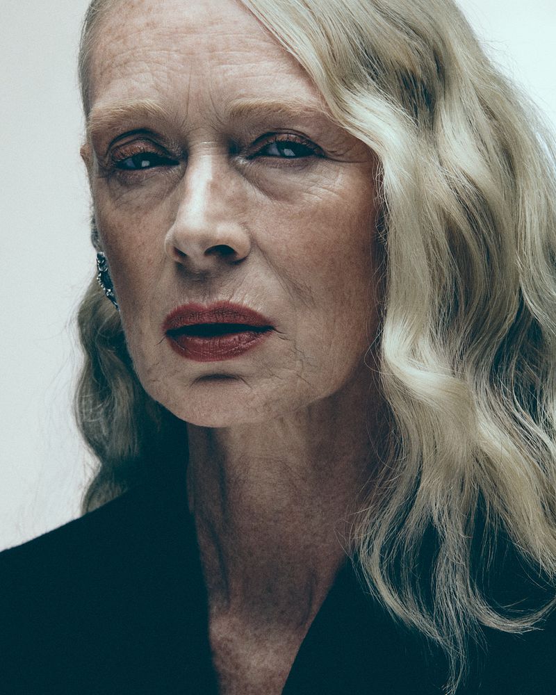 Close-up portrait of a woman with blonde wavy hair, red lipstick, and freckles against a neutral background.