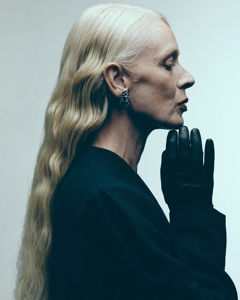 Profile portrait of a blonde woman wearing a black top and leather glove, posing against a pale background