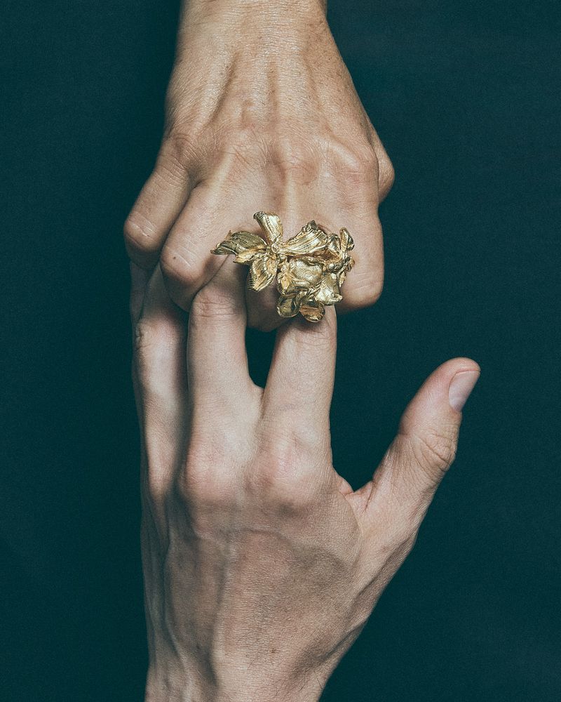 Close-up of two hands holding each other, featuring an ornate gold floral ring worn on one finger against a dark background.
