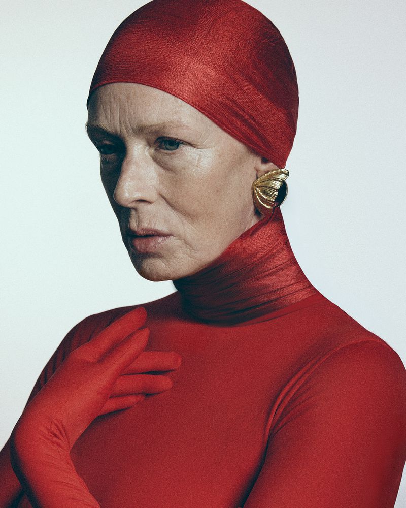 Model in a bright red bodysuit and matching head covering with gold earrings against a light grey background