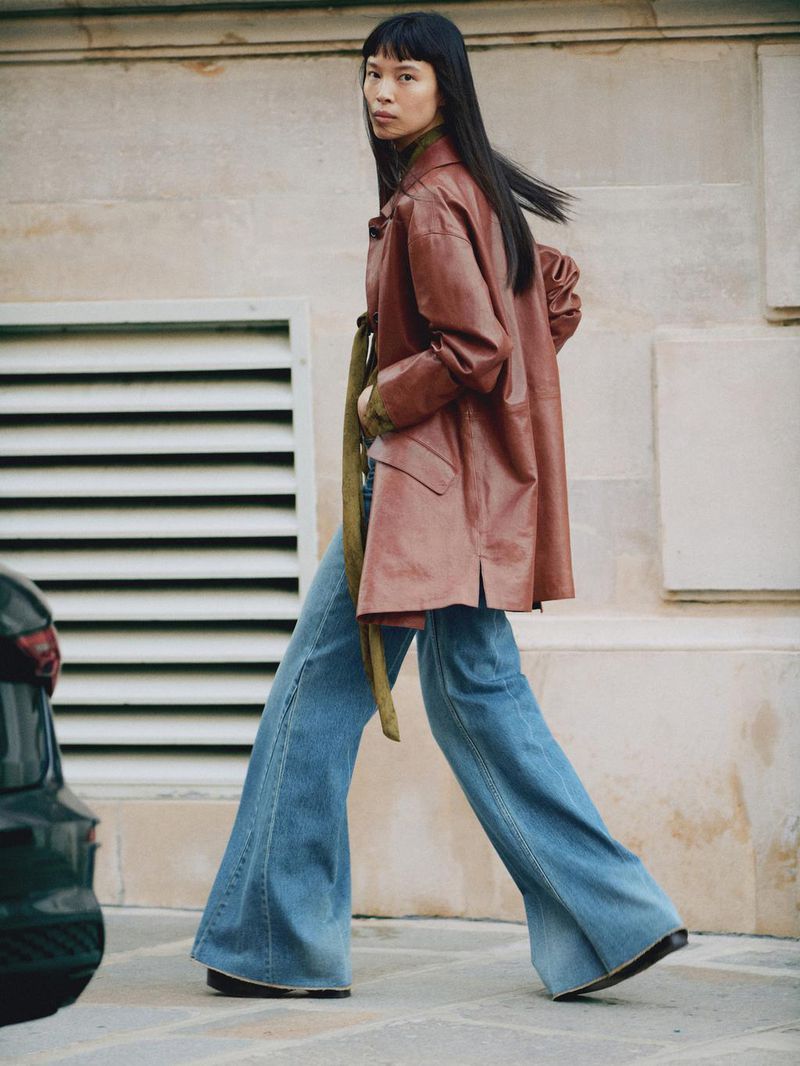 Asian woman in brown leather coat and wide-leg jeans walking on a city street with modern building. Fashion editorial style.
