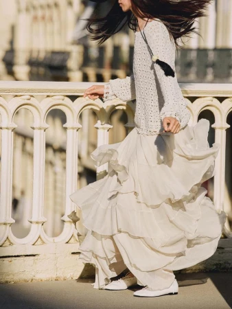 A woman in a long, flowing white skirt and sweater walks on a bridge with an ornate railing, with blurred buildings in the background.