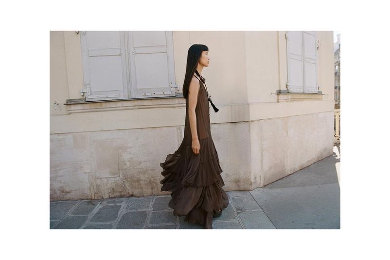 An Asian woman in a long brown dress walks along a city street with white buildings and windows in a fashion or editorial style photo.