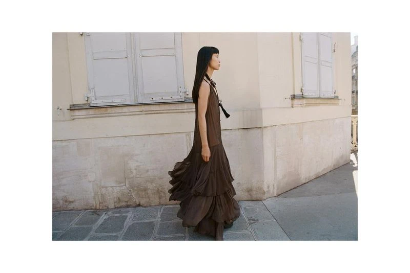 An Asian woman in a long brown dress walks along a city street with white buildings and windows in a fashion or editorial style photo.