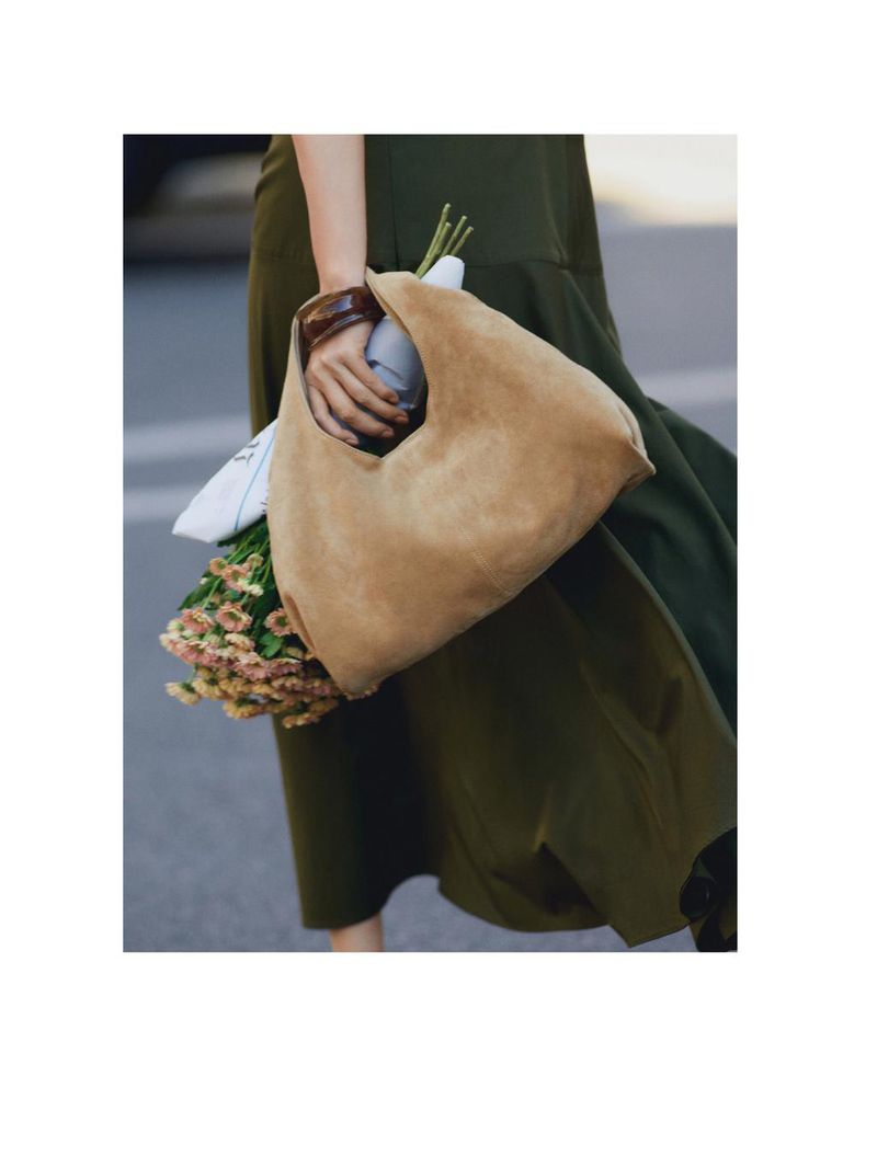 A woman carrying a tan suede handbag and a bouquet of flowers, walking in an olive green dress on a paved street.