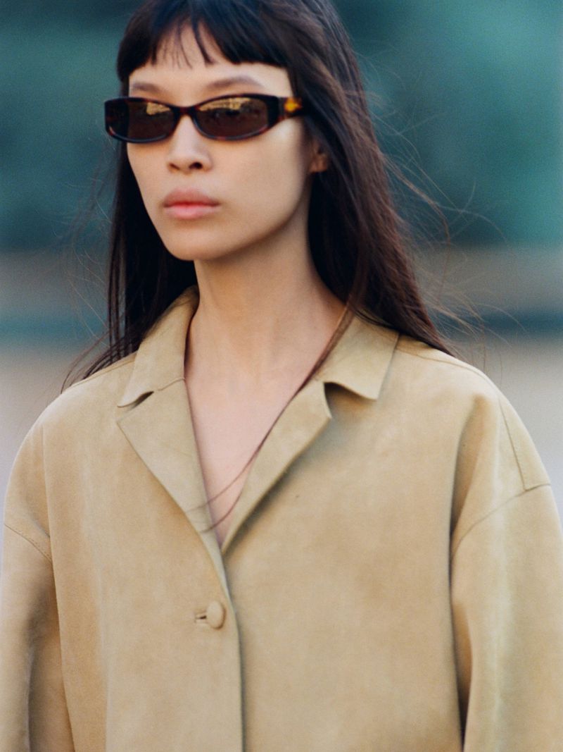 Model Diane Chiu wears dark tortoiseshell sunglasses and a tan suede buttoned coat against an out-of-focus backdrop.