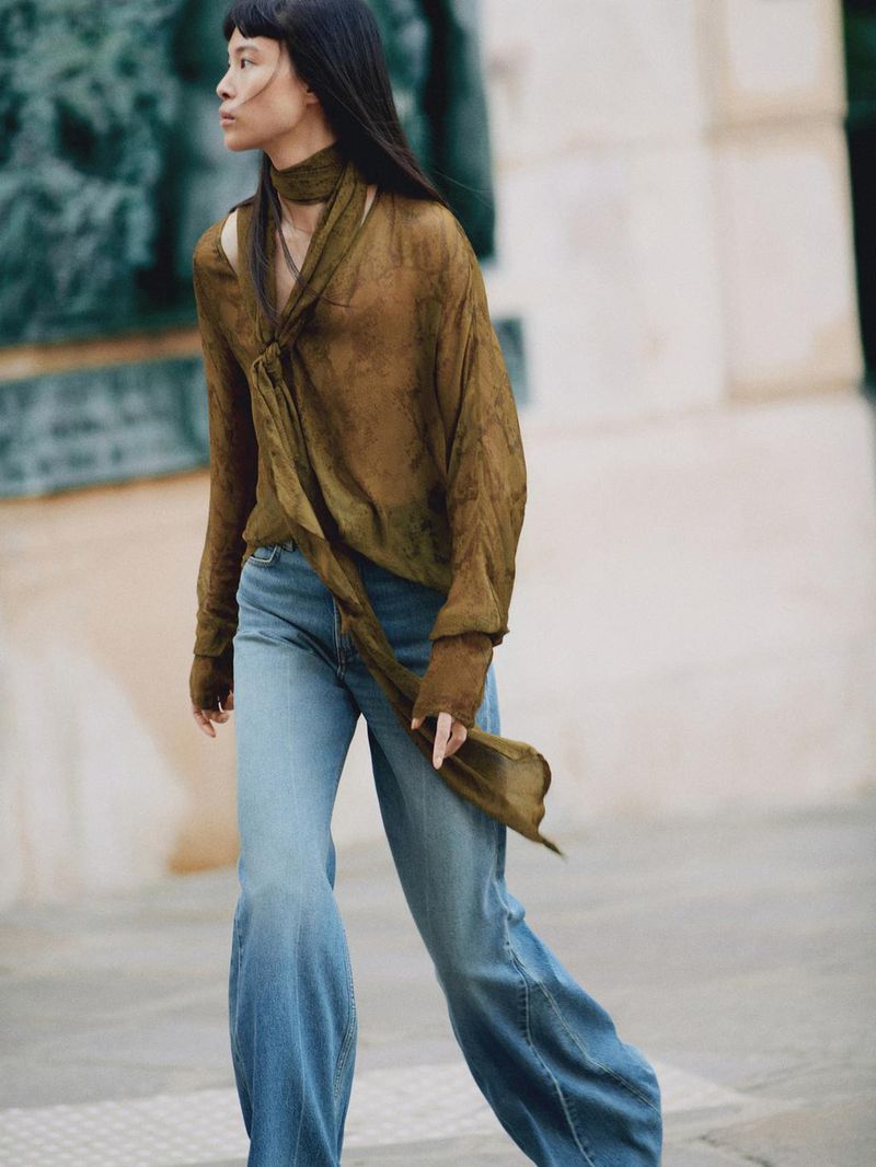 Woman in olive green top and flared jeans walks in an urban setting with neutral tones, showcasing fashion and style.