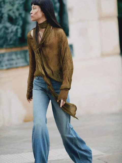 Woman in olive green top and flared jeans walks in an urban setting with neutral tones, showcasing fashion and style.