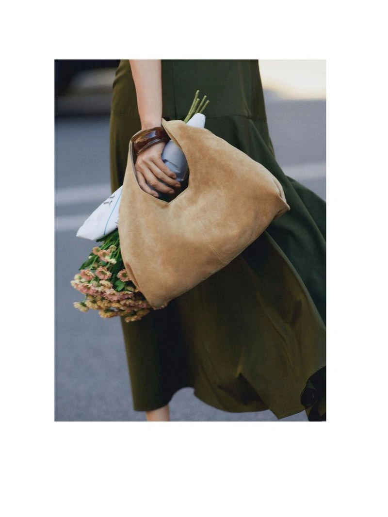 Close-up of a person holding a tan suede tote bag and a bouquet of flowers while wearing an olive green dress on a city street.
