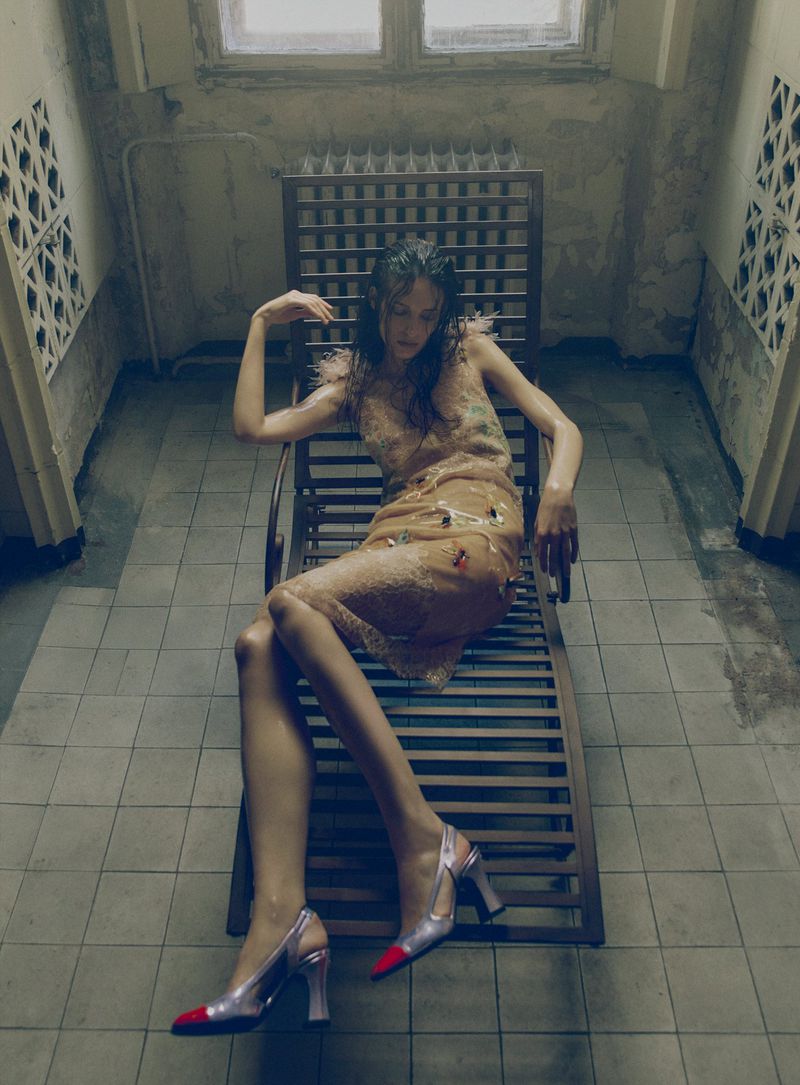 Model seated on a wooden lounger in a tiled room wearing a sheer patterned dress and heels
