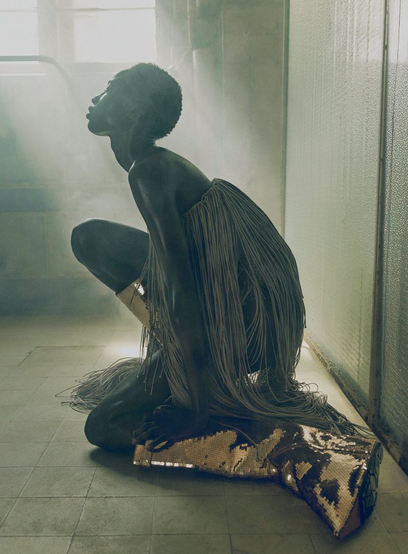 Model kneeling in a hazy industrial space wearing a textured fringe garment and metallic boots