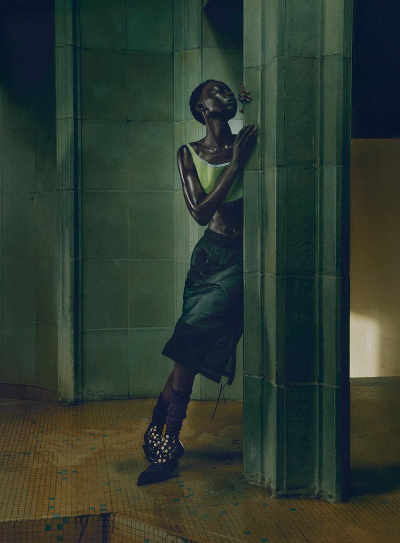 Model leaning against a green tiled column wearing a pale yellow top and dark skirt in a dim architectural interior.