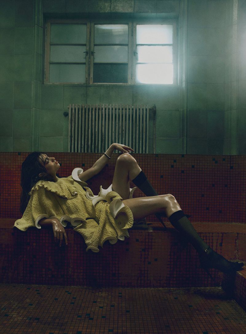 Model in textured yellow ruffled dress reclining in an atmospheric tiled tiled interior with overhead window