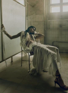 Model seated on a chair in a textured room wearing a textured white top and long skirt