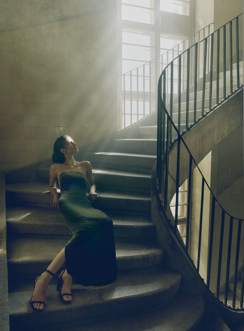 Model in a dark green velvet dress seated on stone stairs beneath a sunlit window for V Magazine