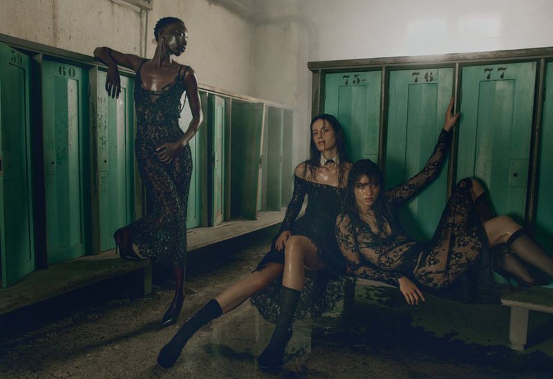 Three models wearing dark lace dresses posed inside a locker room with green lockers for a fashion editorial.