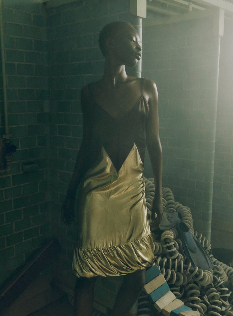 Model wearing a gold and dark slip dress standing against a tiled green wall in hazy lighting.