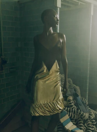 Model wearing a gold and dark slip dress standing against a tiled green wall in hazy lighting.