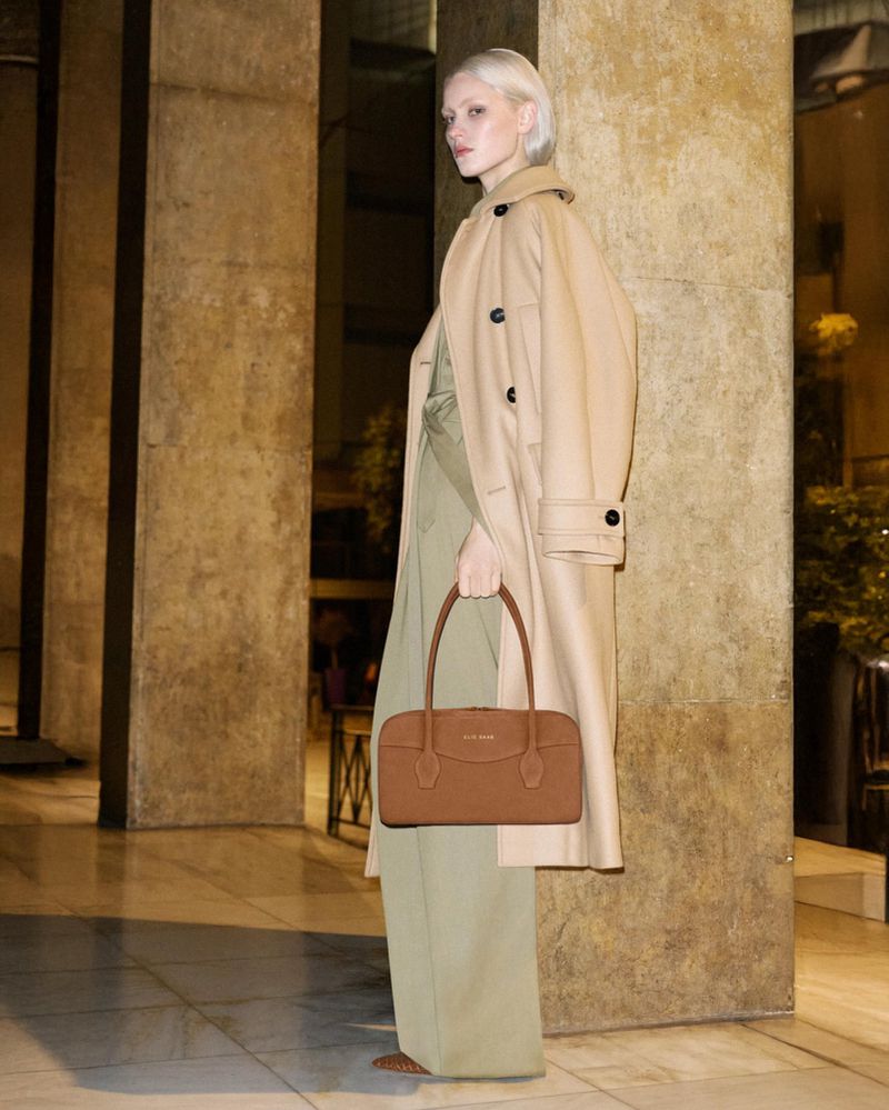 Model with blonde hair wearing a beige coat holding a brown handbag standing against a stone pillar in a hallway