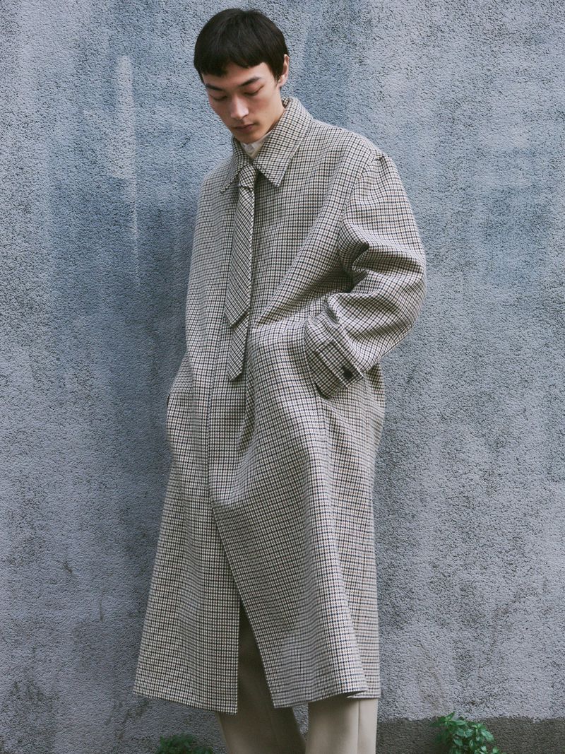 Man wearing a patterned long coat and matching tie standing against a textured grey wall.