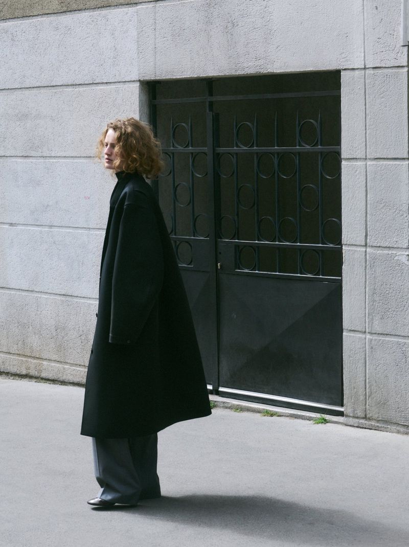 Model with curly hair wearing a long black coat standing outside against a textured concrete wall with a metal gate