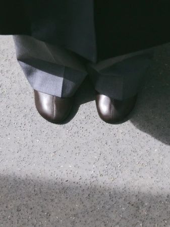 Close-up of brown leather shoes and grey trousers on a concrete ground, photographed from above.