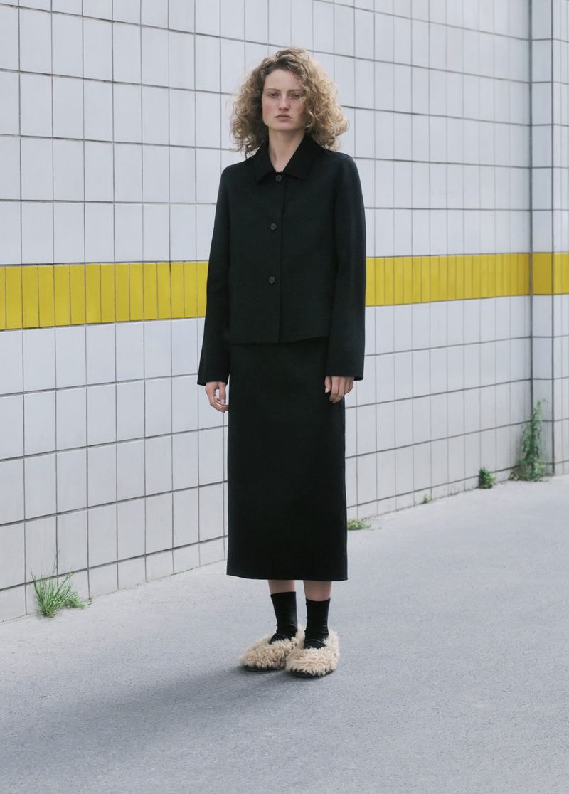 Model standing outdoors wearing a black jacket, matching skirt, black socks, and fluffy textured beige slip-on shoes against a tiled wall