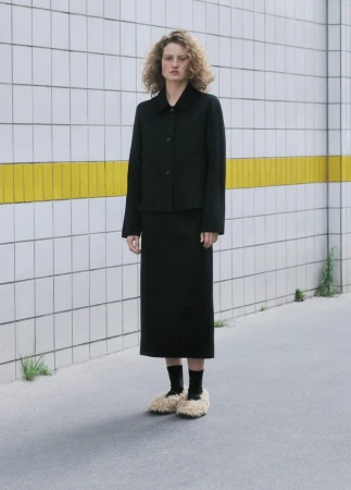 Model standing outdoors wearing a black jacket, matching skirt, black socks, and fluffy textured beige slip-on shoes against a tiled wall
