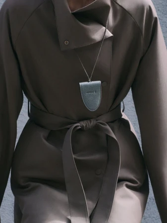 Model wearing a brown belted coat and pendant necklace against a textured wall for ICICLE.