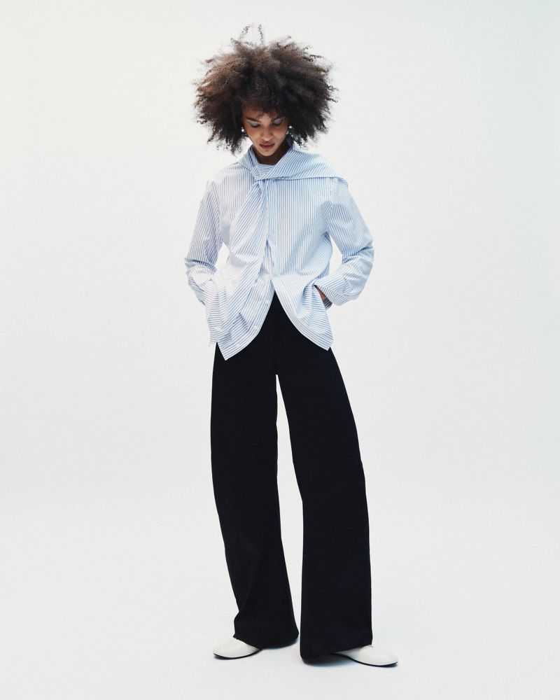 Model with voluminous dark hair wearing a striped blue shirt and black trousers against a plain white background.