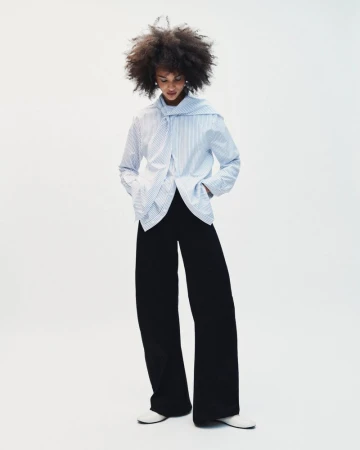 Model with voluminous dark hair wearing a striped blue shirt and black trousers against a plain white background.