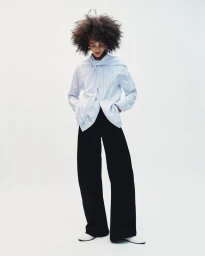 Model with voluminous dark hair wearing a striped blue shirt and black trousers against a plain white background.