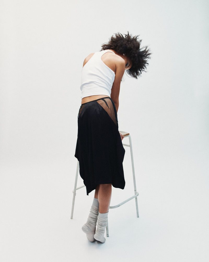 Model with dark curly hair wearing a white tank top and black skirt seated on a stool against a white background