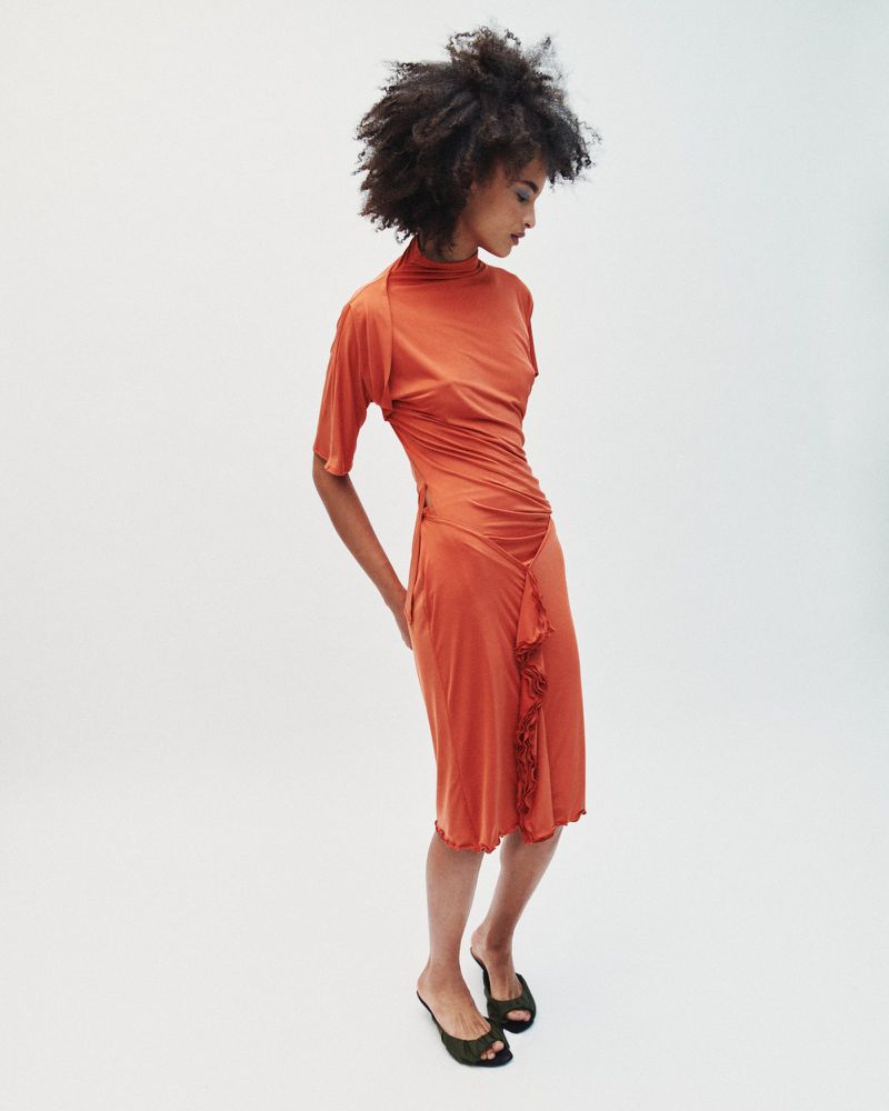 Model with dark curly hair wearing an orange ruched dress against a white studio background