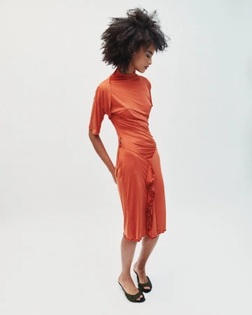 Model with dark curly hair wearing an orange ruched dress against a white studio background