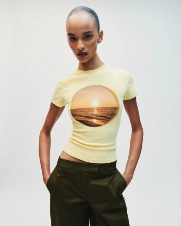 Model wearing a yellow t-shirt with a scenic sunset graphic and dark olive trousers against a plain white background.