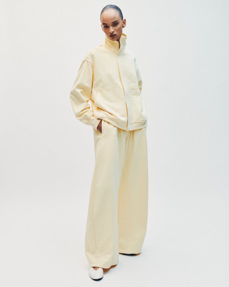Model wearing a pale yellow tracksuit with a high-neck jacket and wide-leg trousers against a white seamless backdrop.